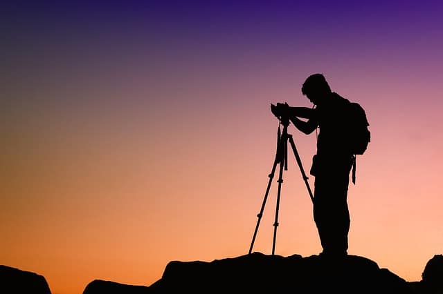 A photographer silhouetted at a tripod on a rock against a sunset sky